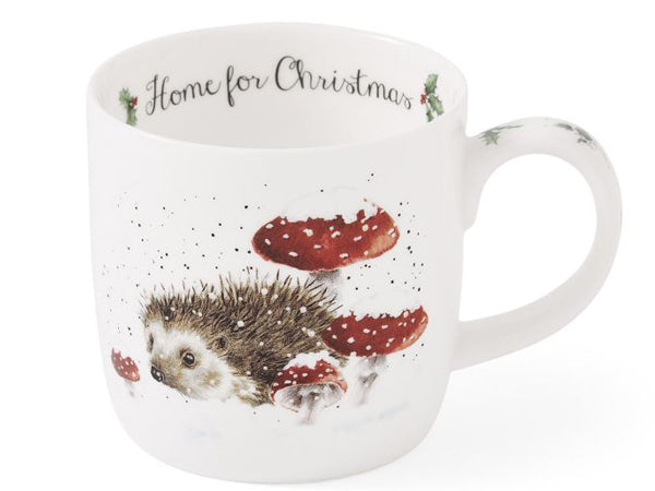 A white ceramic Christmas mug featuring a watercolor illustration of a hedgehog among red-and-white mushrooms, with the text 'Home for Christmas' printed inside the mug.