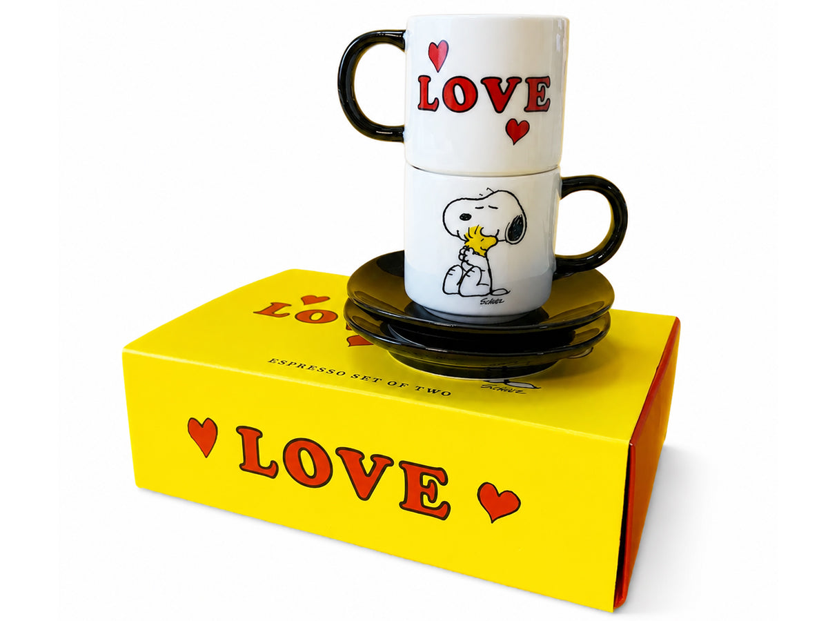 A two-piece espresso mug and saucer set featuring the Peanuts Snoopy and Woodstock design, stacked on top of a bright yellow 'LOVE' branded gift box against a white background.