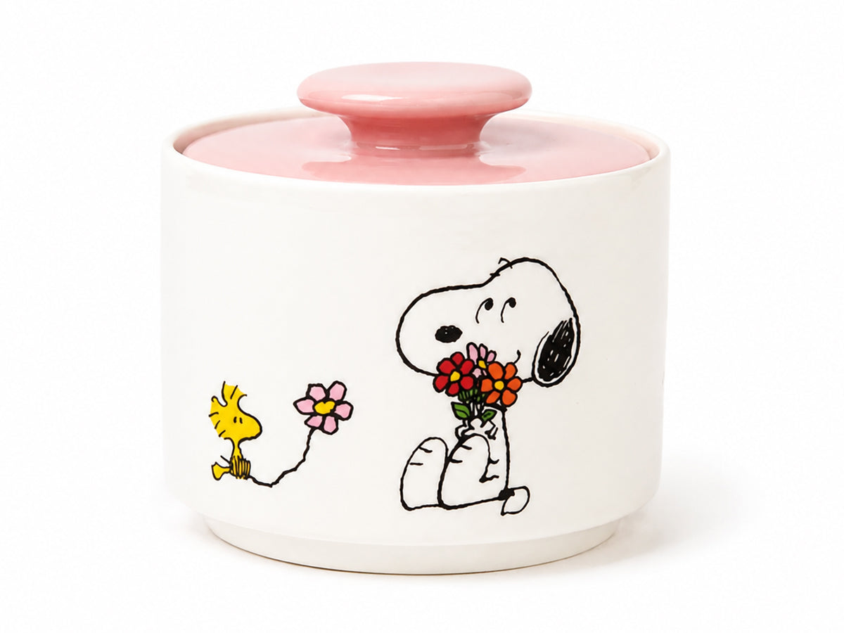 A white ceramic lidded container with a pink lid and a printed illustration of Snoopy holding flowers next to Woodstock, on a white background.