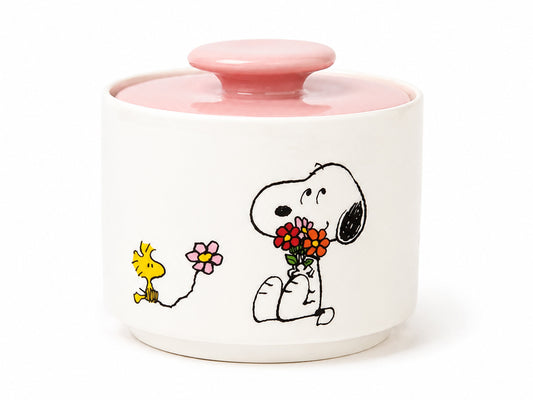 A white ceramic lidded container with a pink lid and a printed illustration of Snoopy holding flowers next to Woodstock, on a white background.