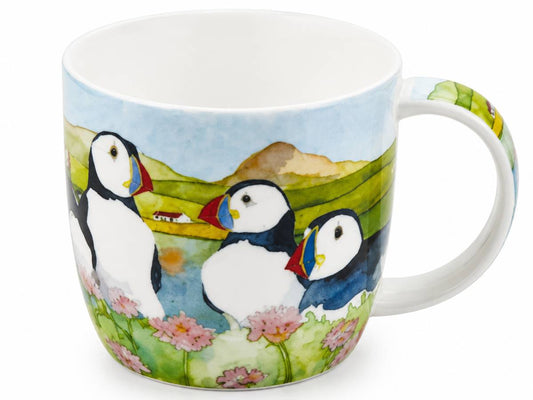A white ceramic mug featuring a colorful watercolor illustration of puffins in a coastal landscape with green hills, pink flowers, and a blue sky.