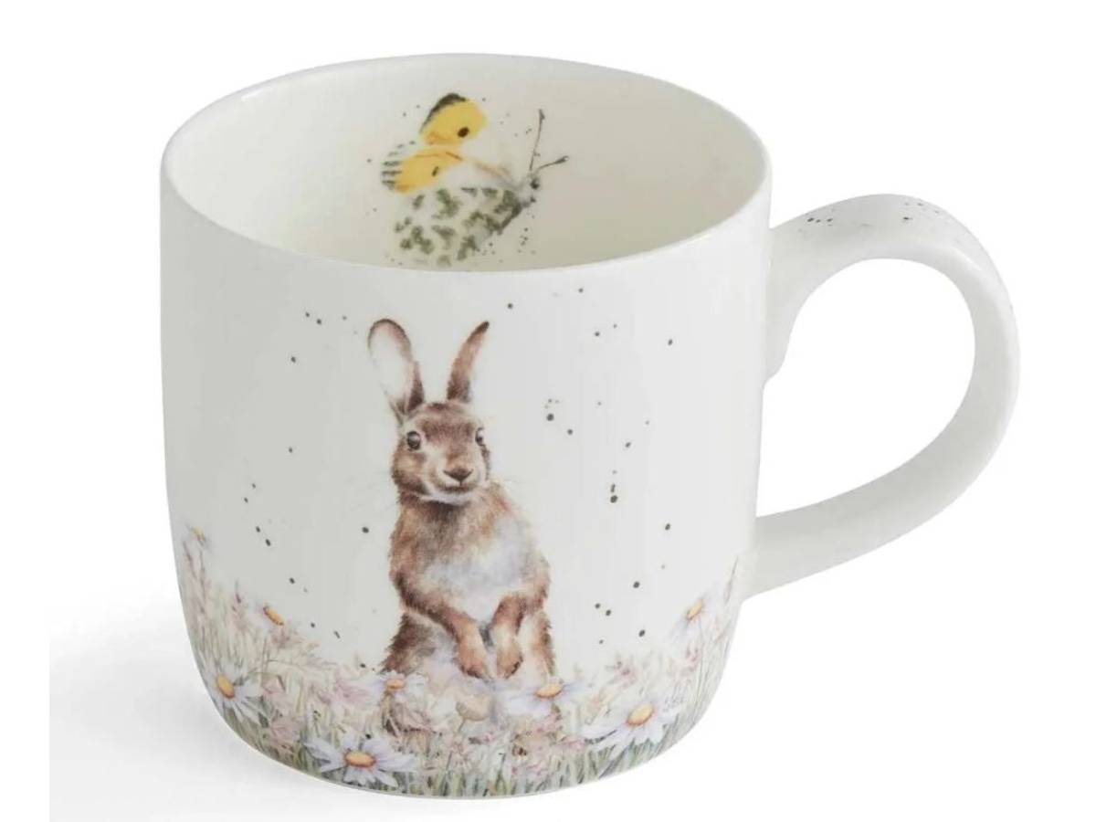 A white ceramic mug featuring a watercolor-style illustration of a brown hare sitting in a field of flowers, with a small yellow bird and greenery printed inside the mug.