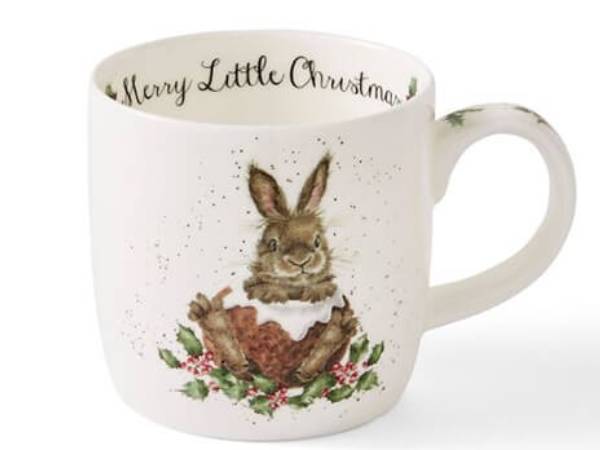 A white ceramic Christmas mug featuring a watercolor illustration of a brown rabbit holding a nutcracker, surrounded by green holly leaves and small pink berries. The inside of the mug is printed with 'Merry Little Christmas' in black script.