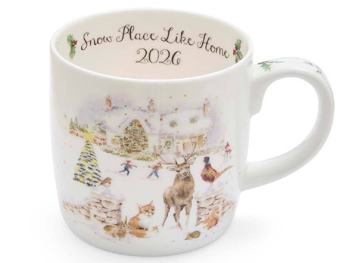 A white ceramic mug featuring a winter village illustration with a deer, dog, hen, and small houses, with the text 'Snow Place Like Home 2026' printed inside the mug.